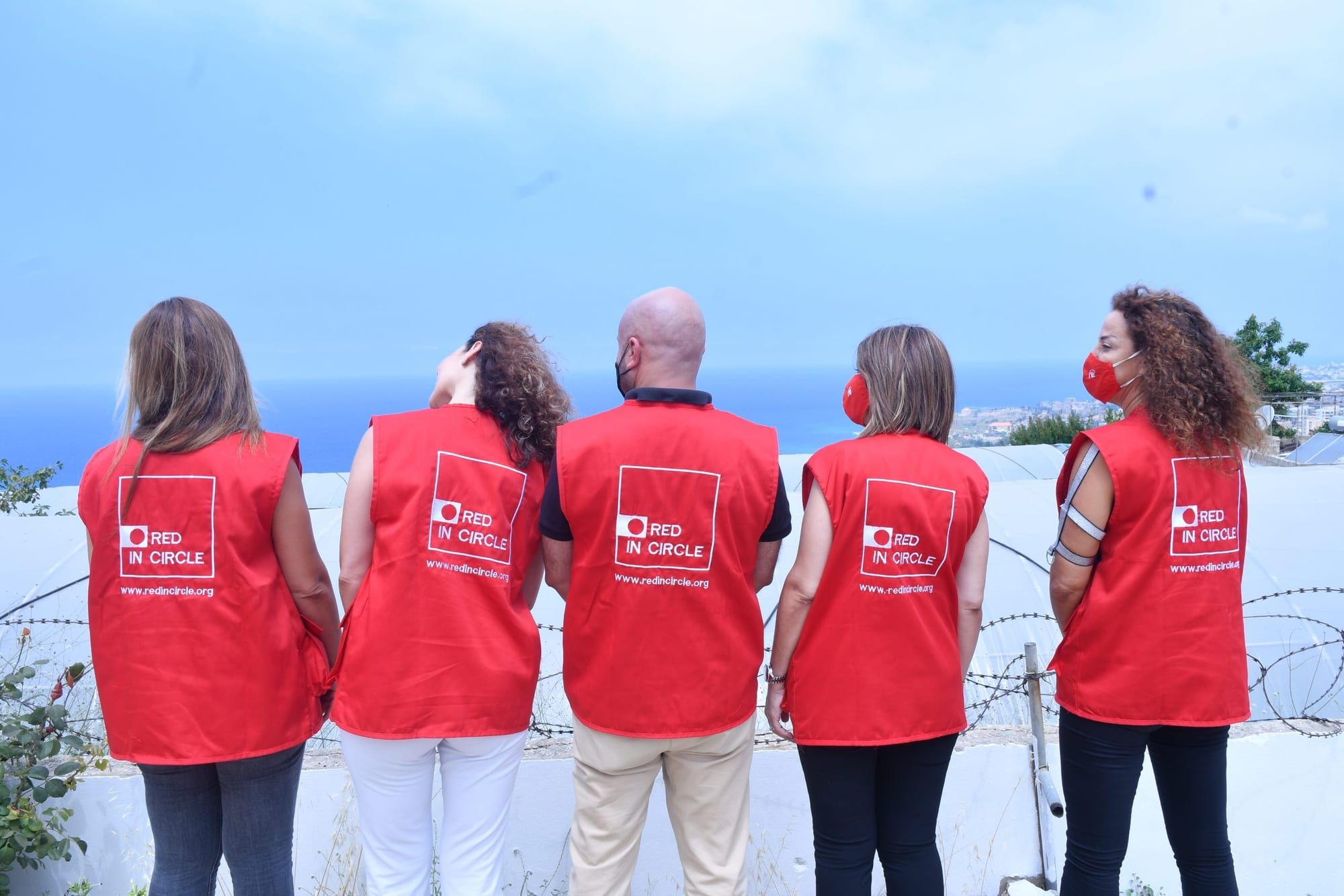 Five Red In Circle volunteers in red vests, seen from behind, looking out over the Lebanese coast.